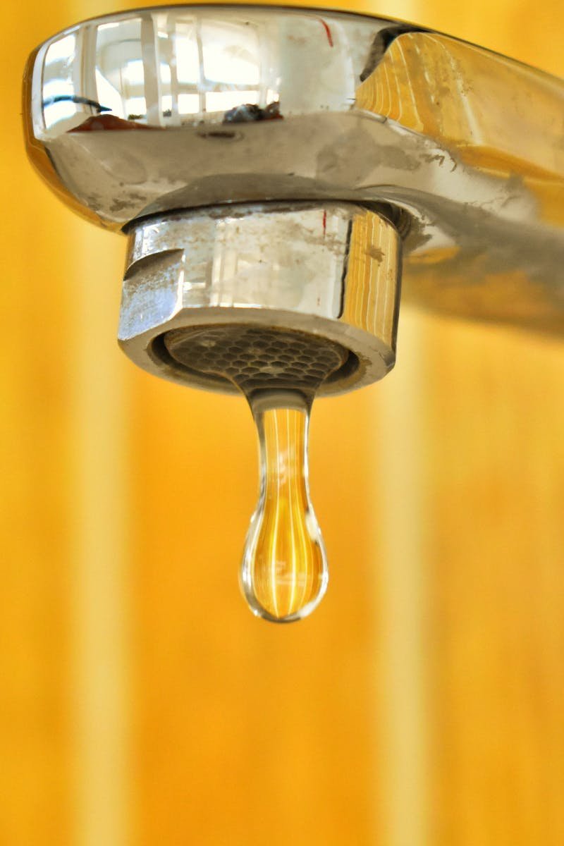 A close-up shot of a water droplet hanging from a stainless steel faucet, highlighting surface tension.